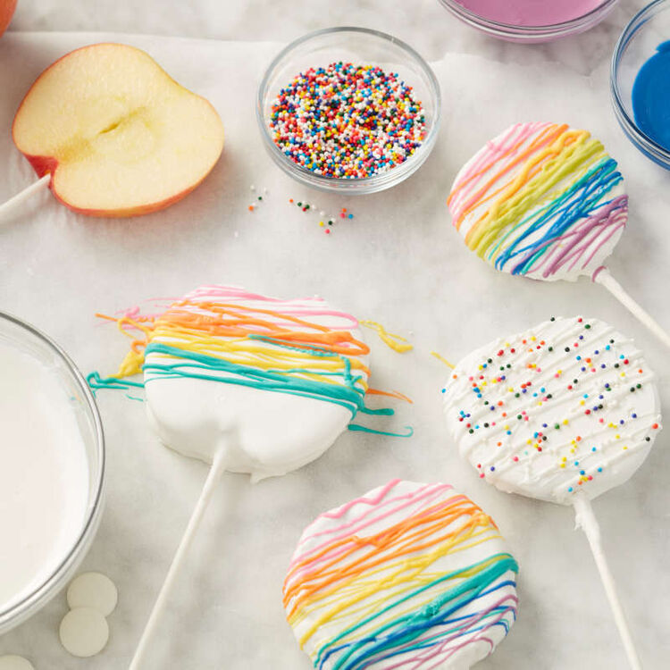 White Candy Melt covered apple slices on a stick drizzled with green, yellow, and red Candy Melts